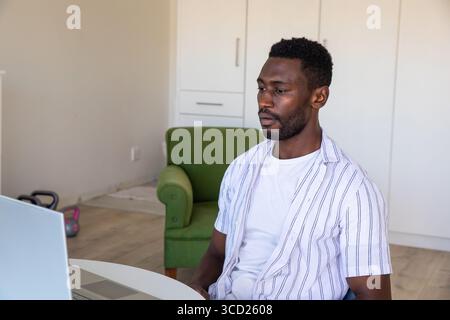 Uomo afroamericano che lavora su un computer portatile nell'area soggiorno di casa vicino a kettlebell e poltrona, spazio fotocopie Foto Stock