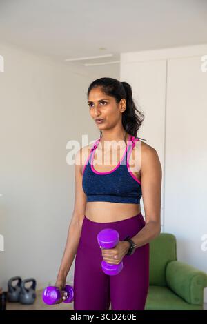 Donna indiana con manubri viola che indossa reggiseno sportivo e leggings nel soggiorno vicino ai kettlebell Foto Stock