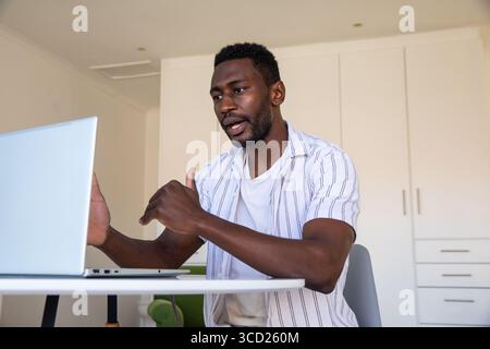Uomo afroamericano seduto al tavolo rotondo in appartamento che gestiva una riunione online con un computer portatile Foto Stock