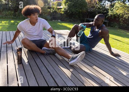 Diversi amici maschi che riposano sul ponte di legno nel cortile posteriore con abbigliamento sportivo e che tengono bottiglie d'acqua Foto Stock
