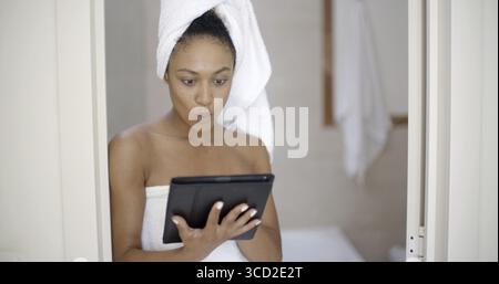 Attraente giovane donna nera con un tablet touchscreen appoggiato sulla porta del bagno Foto Stock