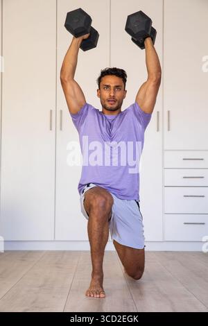 Uomo asiatico che esegue un affondo a forma di manubrio sopra la testa nella palestra domestica sul pavimento indossando camicia viola, pantaloncini grigi Foto Stock