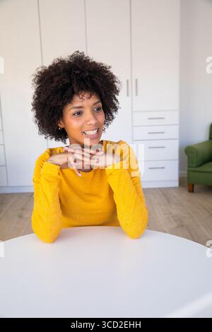 Donna afro-americana seduta a un tavolo bianco in camera con piano di senape in armadietti incorporati Foto Stock