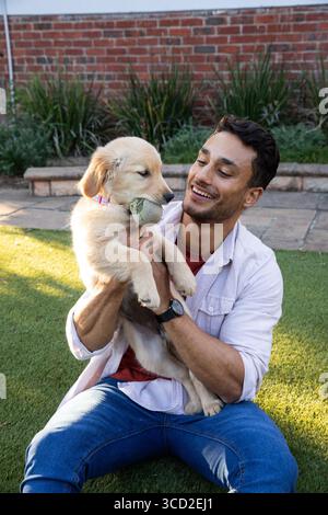 Uomo seduto a gambe incrociate sul prato del cortile che tiene il cucciolo Golden retriever con la palla da tennis Foto Stock