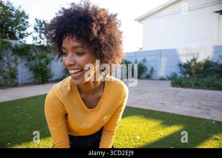 Donna afro-americana che si piega ridendo nel cortile posteriore indossando un top giallo senape e una semplice collana Foto Stock