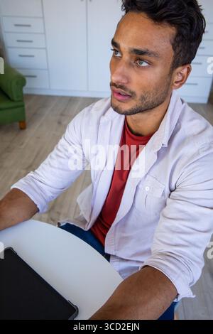 Uomo indiano che lavora su un tablet a un tavolo rotondo bianco nella zona giorno della casa con poltrona verde Foto Stock