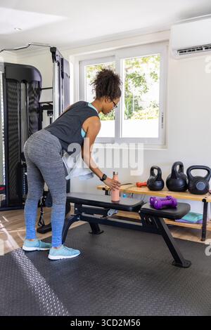 Donna afroamericana che si piega sulla panchina in palestra per raggiungere una bottiglia d'acqua rosa e uno spazio fotocopie Foto Stock