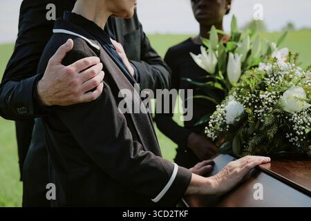 Donna caucasica di mezza età che viene confortata da un uomo caucasico di mezza età mentre si trova accanto alla bara con donna nera che tiene il bouquet funebre durante la cerimonia di sepoltura all'aperto Foto Stock
