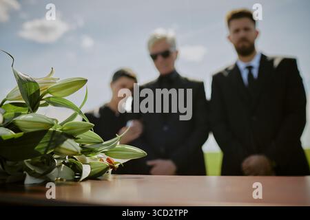Tre adulti caucasici in piedi insieme vicino alla bara, due uomini e una donna vestiti con abiti scuri, donna appoggiata su un uomo più anziano, bouquet di gigli appoggiati su una bara in primo piano Foto Stock