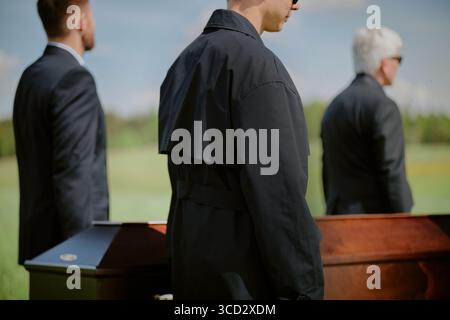 Tre uomini, tra cui un giovane uomo caucasico adulto, un uomo caucasico di mezza età, un anziano uomo caucasico in piedi accanto a una bara di legno all'aperto durante la cerimonia funebre di giorno Foto Stock