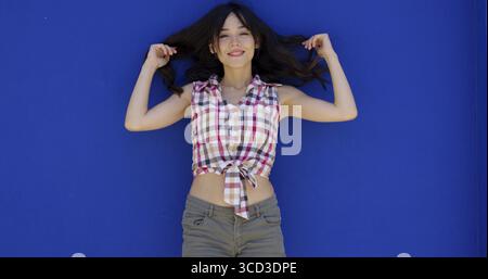 Giovane donna sorridente e felice che tiene i suoi bei lunghi capelli scuri tra le mani su uno sfondo blu che strizza alla telecamera Foto Stock