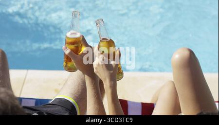 Coppia rilassante in piscina con birre che si godono le vacanze estive vista ravvicinata delle loro mani e delle bottiglie piene Foto Stock