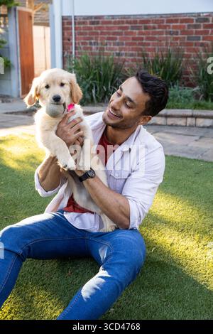 Uomo seduto nel prato del cortile che tiene in mano un cucciolo Golden retriever con colletto rosa e palla da tennis Foto Stock