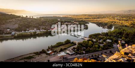 Vista panoramica di un fiume che si snoda attraverso una città con un ponte, circondato da vegetazione lussureggiante e montagne sullo sfondo durante il tramonto. Foto Stock