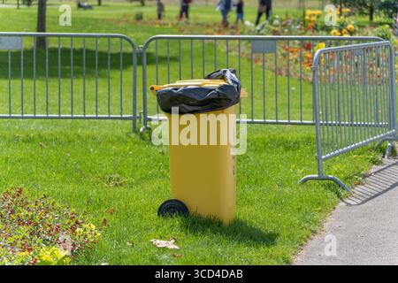 Un cestino giallo brillante con un sacchetto nero si trova su un prato erboso, recintato da barriere metalliche. Fiori e persone appaiono sullo sfondo. Foto Stock