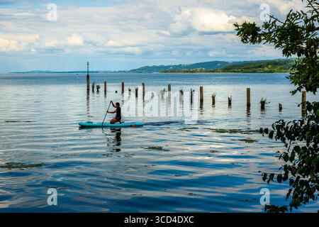 Yverdon les Bains, Svizzera - 24 luglio 2025: Una donna in paddleboard su un lago calmo di Neuchatel con riflessi sull'acqua, circondato da greene Foto Stock