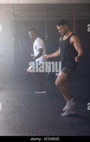 Diversi amici maschi che saltano la corda in palestra su un tappeto da squat rack con cinturino rosso Foto Stock