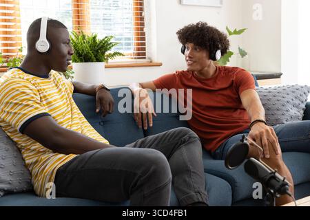 Diversi amici maschi registrano podcast su un divano blu nel soggiorno con microfono e cuffie Foto Stock