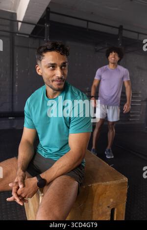 Amici maschi diversi che riposano su una scatola plyometrica in legno in palestra accanto a un rack per squat e a uno spazio per copiare Foto Stock