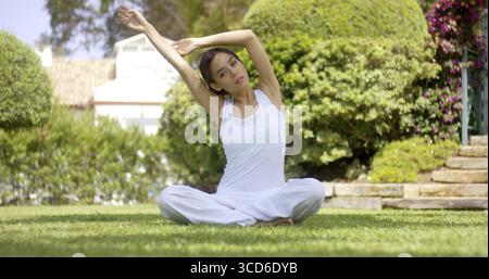 Splendida e tranquilla giovane donna in bianco seduta su un prato con le braccia allungate ai lati Foto Stock