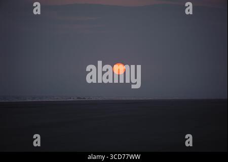 Un sole infuocato tramonta su una spiaggia tranquilla, dipingendo il cielo con sfumature di arancione e viola. Tranquilla scena costiera. Foto Stock