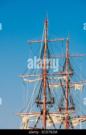 Una maestosa nave alta si erge orgogliosamente con i suoi alberi e le intricate attrezzature contro un cielo blu brillante. Le vele si appendono dolcemente mentre l'imbarcazione attende il suo nex Foto Stock