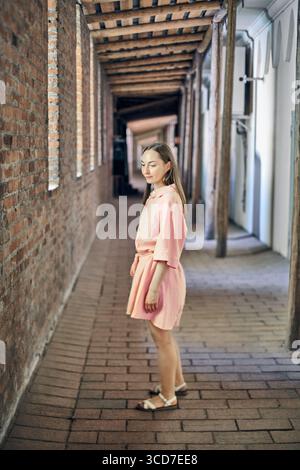 Giovane donna in abito rosa si erge in un corridoio figo. Mattoni rustici e travi di legno accentuano il fascino. La luce naturale soffusa viene filtrata, migliorando seren Foto Stock
