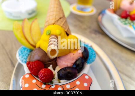 Lindau, Baviera, Germania - 11 agosto 2025: Coppa estiva per bambini: Pinocchio di gelato con cappello e naso arrotolato, decorato con fragole, uva e fette di mele. Dolce piacere nella gelateria *** Sommerlicher Kinderbecher: Pinocchio aus Eis mit Waffelhut und Röllchennase, dekoriert mit Erdbeeren, Trauben und Apfelspalten. Süßer Genuss in der Eisdiele Foto Stock