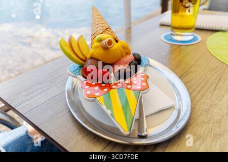 Lindau, Baviera, Germania - 11 agosto 2025: Coppa estiva per bambini: Pinocchio di gelato con cappello e naso arrotolato, decorato con fragole, uva e fette di mele. Dolce piacere nella gelateria *** Sommerlicher Kinderbecher: Pinocchio aus Eis mit Waffelhut und Röllchennase, dekoriert mit Erdbeeren, Trauben und Apfelspalten. Süßer Genuss in der Eisdiele Foto Stock
