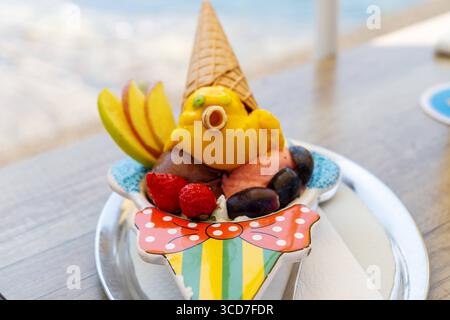 Lindau, Baviera, Germania - 11 agosto 2025: Coppa estiva per bambini: Pinocchio di gelato con cappello e naso arrotolato, decorato con fragole, uva e fette di mele. Dolce piacere nella gelateria *** Sommerlicher Kinderbecher: Pinocchio aus Eis mit Waffelhut und Röllchennase, dekoriert mit Erdbeeren, Trauben und Apfelspalten. Süßer Genuss in der Eisdiele Foto Stock