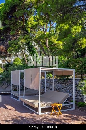 Lussuosa zona lounge esterna con moderni lettini a baldacchino su una terrazza in legno nel giardino dell'hotel, circondata da lussureggianti pini in una giornata di sole. Moderno ed elegante Foto Stock