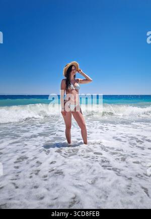 Bella donna che posa in bikini e cappello di paglia, si erge tra le onde spumeggianti del mare azzurro, sotto un cielo estivo senza nuvole. Vacanze estive Foto Stock