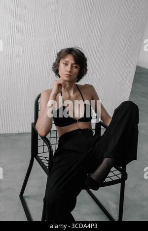 il modello è caratterizzato da una posa accattivante con un top bikini nero e pantaloni morbidi, seduti su una sedia elegante in uno spazio luminoso e minimalista. Li naturali Foto Stock