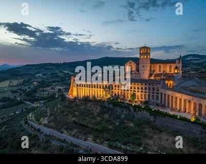La vista aerea della Basilica di San Francesco d'Assisi si illumina di luce dorata contro il crepuscolo blu profondo, Assisi, Umbria, Italia. Foto Stock