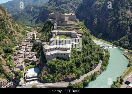 Vista aerea dell'imponente fortezza di Fort Bard arroccata in cima a una collina verdeggiante, che si affaccia sul tortuoso fiume Dora Baltea e sul pittoresco villaggio sottostante, Bard, Valle d'Aosta, Italia. Foto Stock