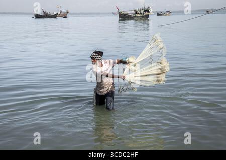 Cox's Bazar, Bangladesh - 29 novembre 2022: Veduta di un pescatore in vita nelle tranquille acque ondulate, che gettano una rete, con barche da pesca che si stagliano all'orizzonte. Foto Stock
