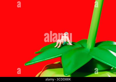 un uomo in miniatura con costume da bagno e cappuccio si siede dietro con le braccia allungate su una foglia verde di plastica posta sopra una bottiglia a forma di mora verde, Foto Stock