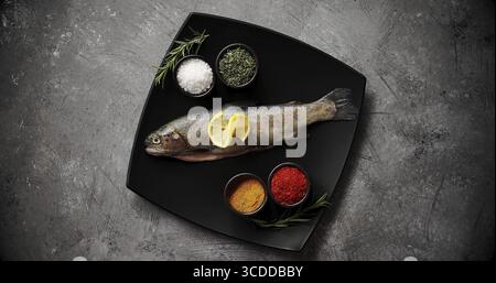 Da sopra il colpo di piastra in ceramica con assortimento di spezie aromatiche fresche e trota cruda permanente sulla superficie del tavolo di pietra Foto Stock