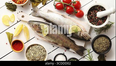 Da sopra il colpo di varie spezie aromatiche e i pomodori maturi che giace attorno al bordo di taglio con crudo fresco trota su legno bianco il piano portapaziente Foto Stock