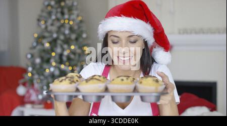 Giovane donna sorridente con un cappello di Babbo Natale rosso in piedi di fronte a un albero di Natale che regge un vassoio di cupcake appena sfornati per celebrare la festa Foto Stock