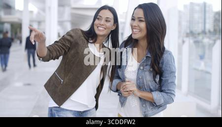 Giovane donna attraente e alla moda che punta a qualcosa mentre attira l'attenzione della sua compagna femminile su di esso entrambe le donne sono sorridenti e felici urban es Foto Stock