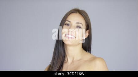 Una splendida giovane donna adulta dai lunghi capelli castani con un piacevole sorriso su sfondo grigio Foto Stock
