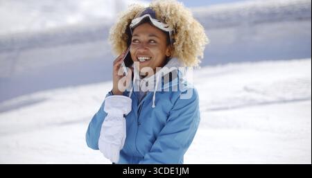 Donna single con occhiali da neve blu e guanto bianco che ride con il telefono all'orecchio e la neve brillante alle spalle Foto Stock