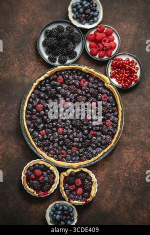 Torta rotonda fresca fatta in casa con frutti di bosco, lamponi, more, gelatina di ribes rosso e nero. Vista dall'alto, piatto su tavolo di rustit scuro Foto Stock