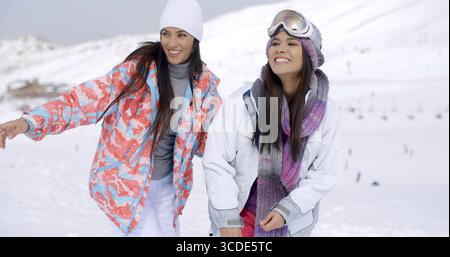 Due graziose giovani amiche in una stazione sciistica di montagna che si affacciano lateralmente sulla cornice in un paesaggio innevato e una pista da sci in lontananza con c Foto Stock