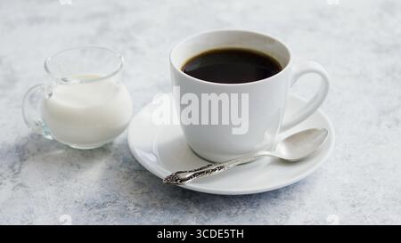 Disposizione di una caraffa di vetro piccola con latte fresco su un tavolo in marmo con una tazza di caffè bianca e un cucchiaio d'argento sul piattino Foto Stock