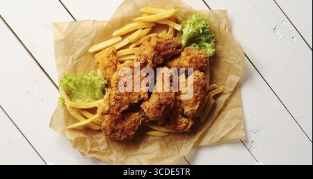 Da sopra il colpo di foglie di lattuga fresca e golosa patatine fritte che giace vicino al mucchio di deliziose Alette di pollo fritte sul legname bianco il piano portapaziente Foto Stock