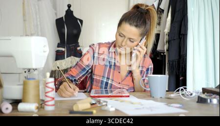 Giovane donna in abbigliamento casual che parla al telefono mentre disegna schizzi e usa un tablet in salotto Foto Stock