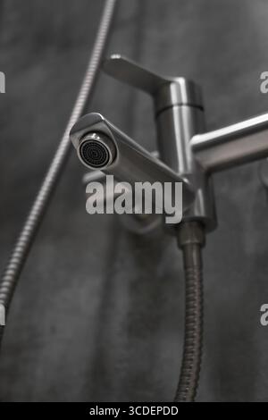 Primo piano di un rubinetto doccia in acciaio inossidabile con tubo flessibile in metallo, montato su una parete piastrellata grigia in un bagno contemporaneo, con un moderno piumone Foto Stock
