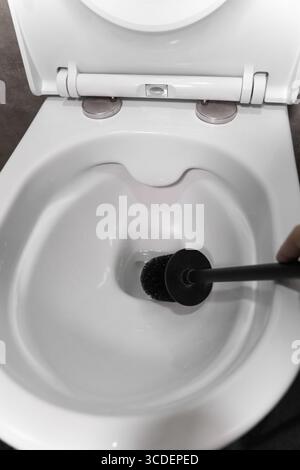 Lo spazzolino per WC nero che si libra sopra una ciotola bianca aperta e immacolata, sottolineando l'importanza dell'igiene del bagno e promuovendo un senso di pulizia Foto Stock
